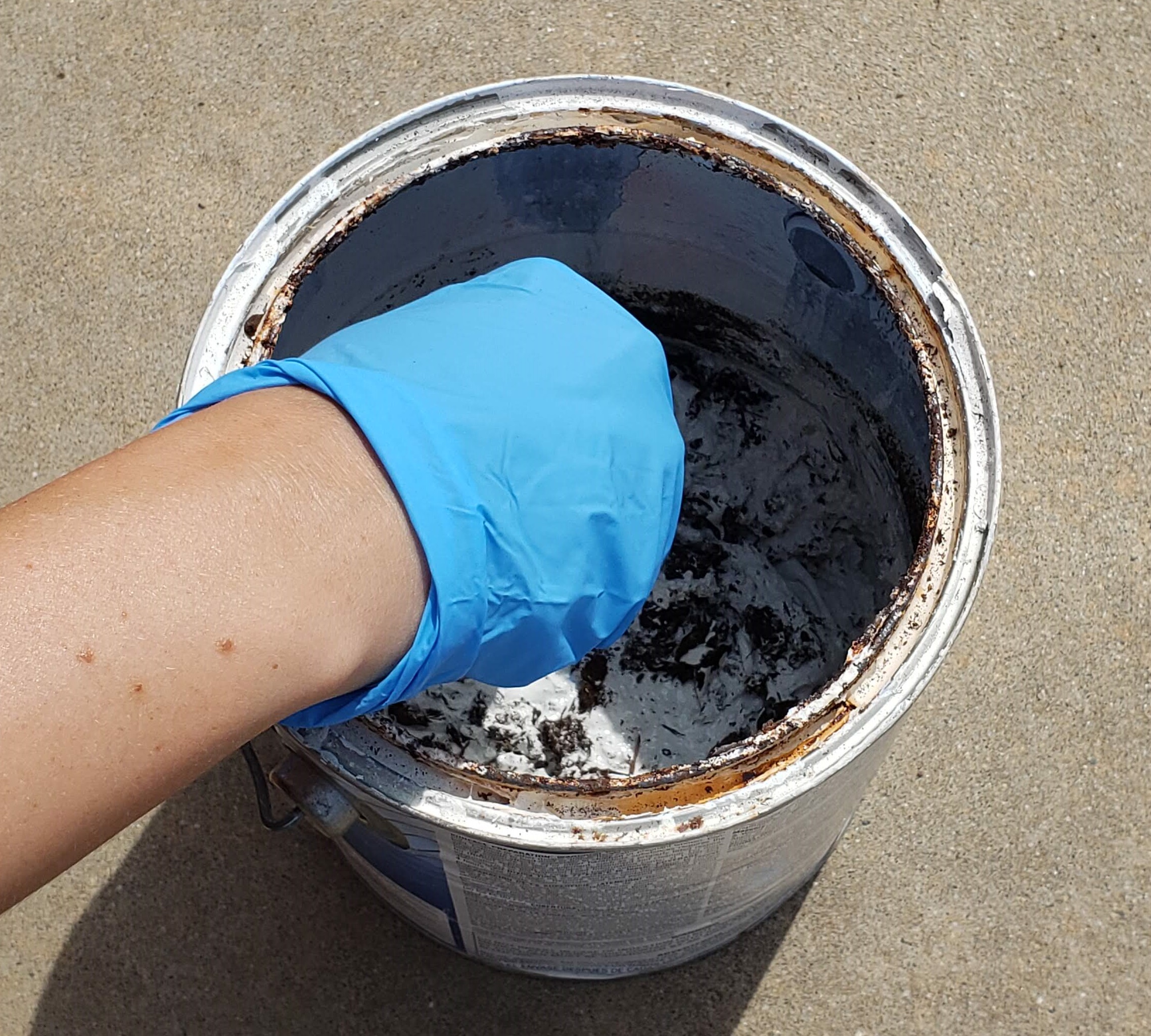 Hand using a small cylindrical tool to mix the dirt and paint in the 1 gallon paint can