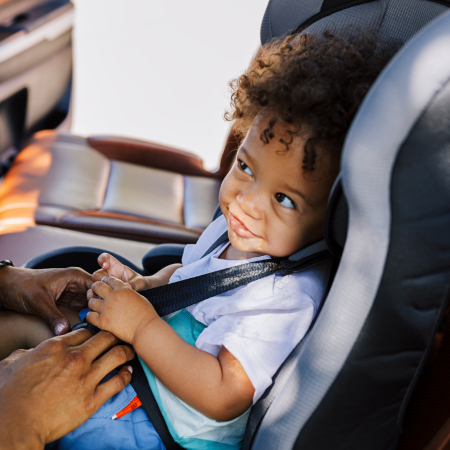 A little girl with curly hair is being buckled into a car seat by an adult off-camera.