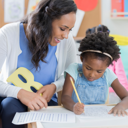 A small Black child writes on a page while being helped by an adult teacher in a classroom.