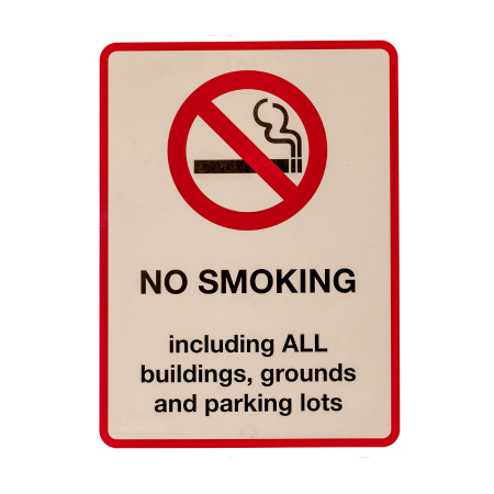 A rectangular vertical white sign with red trim around the outside. At the top of the sign is a cartoon drawing of a cigarette with a big red circle and diagonal line over it, with the words "NO SMOKING" in big letters below it above, including ALL buildings, grounds, and parking lots.