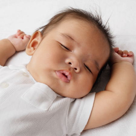 A baby sleeps in a white onesie with their arms by their head on a white sheet.