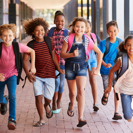 A group of children dressed for school and wearing backpacks runs down an external hallway at a school.