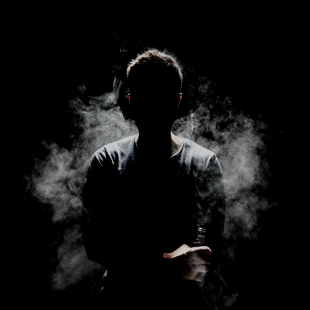 An individual is in darkness and mostly in black while vape smoke floats around them.