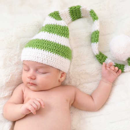 A baby lies on their back on a blanket with a knit cap on their head with a long tail